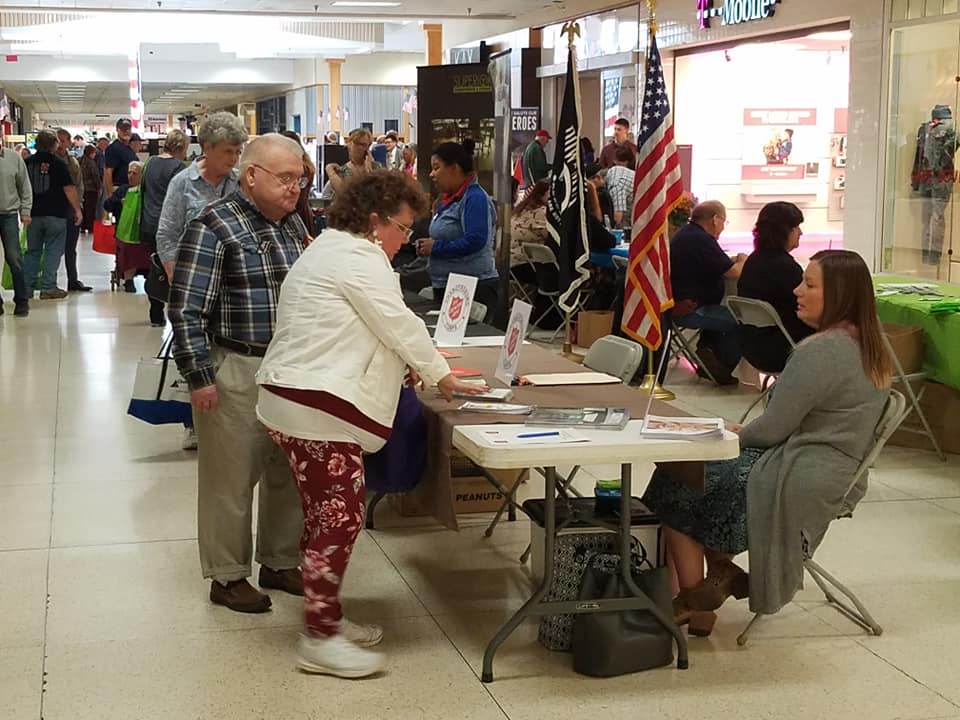 People interact at tables in a busy indoor event space, with informational displays and an American flag visible.