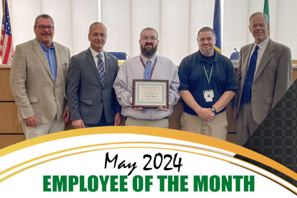 Five men standing in an office; one holds a framed certificate. Text reads, "May 2024 Employee of the Month.