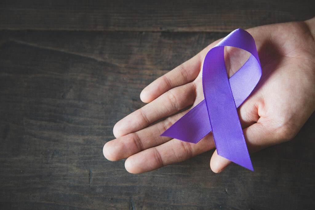 A hand holds a purple awareness ribbon resting on a wooden surface.