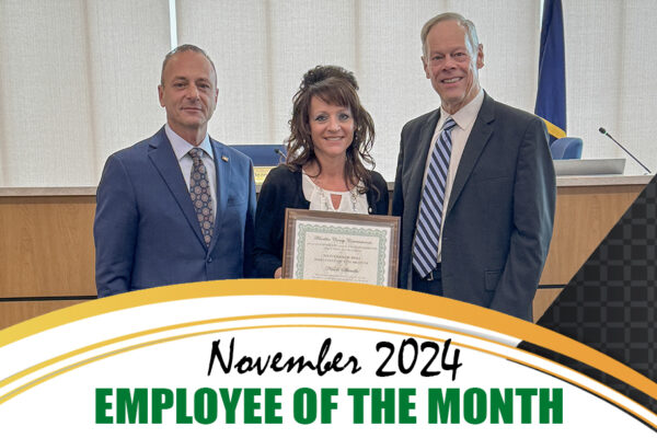 Three individuals stand together, one woman holding a certificate. Text reads "November 2024 Employee of the Month." Two men are on either side, wearing suits.